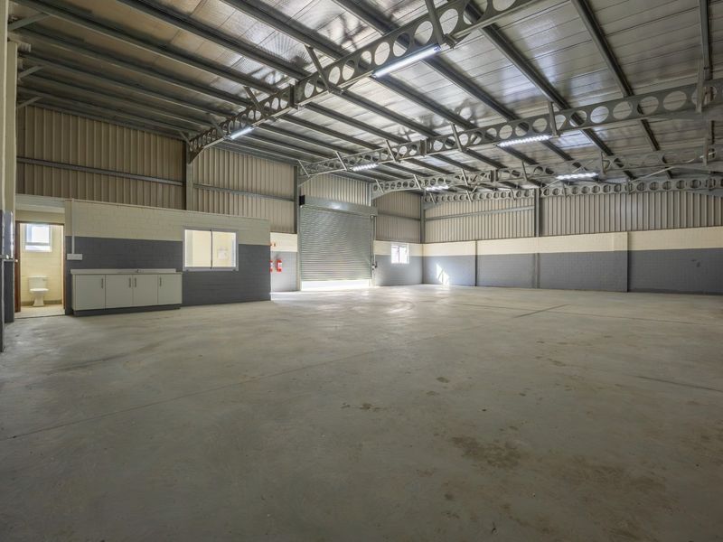 Spacious industrial warehouse unit featuring high ceilings with exposed steel trusses, concrete flooring, a roller shutter door, and an integrated office or utility area.