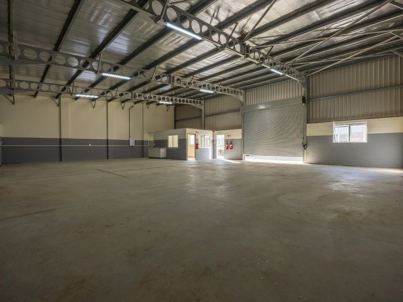 Spacious industrial warehouse unit featuring high ceilings with exposed steel trusses, concrete flooring, a large roller shutter door, and an integrated internal office space.