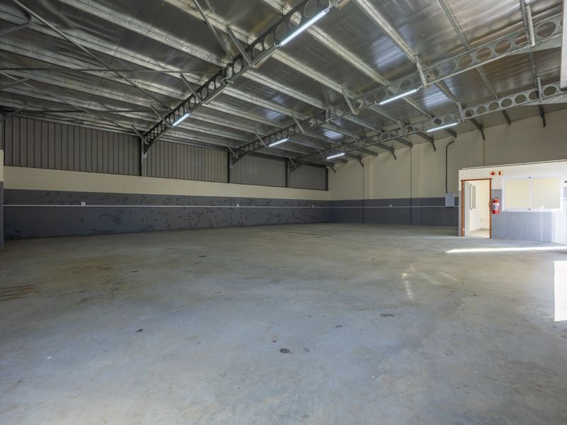 Spacious industrial warehouse unit featuring high exposed steel roof trusses, concrete flooring, a roller shutter door, and an integrated internal office space.