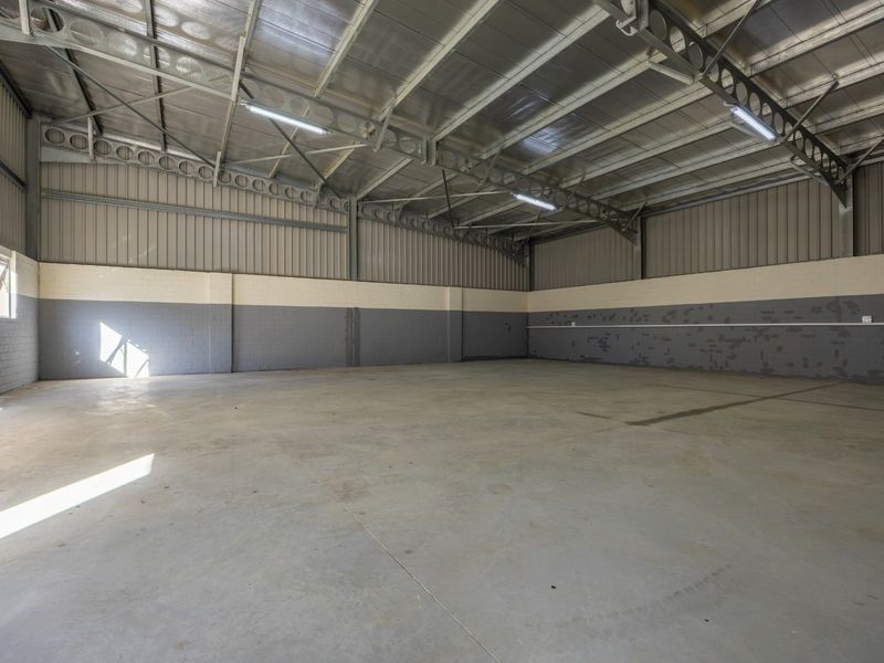 A spacious, empty industrial warehouse unit featuring high ceilings with exposed steel trusses, concrete flooring, and painted blockwork walls.