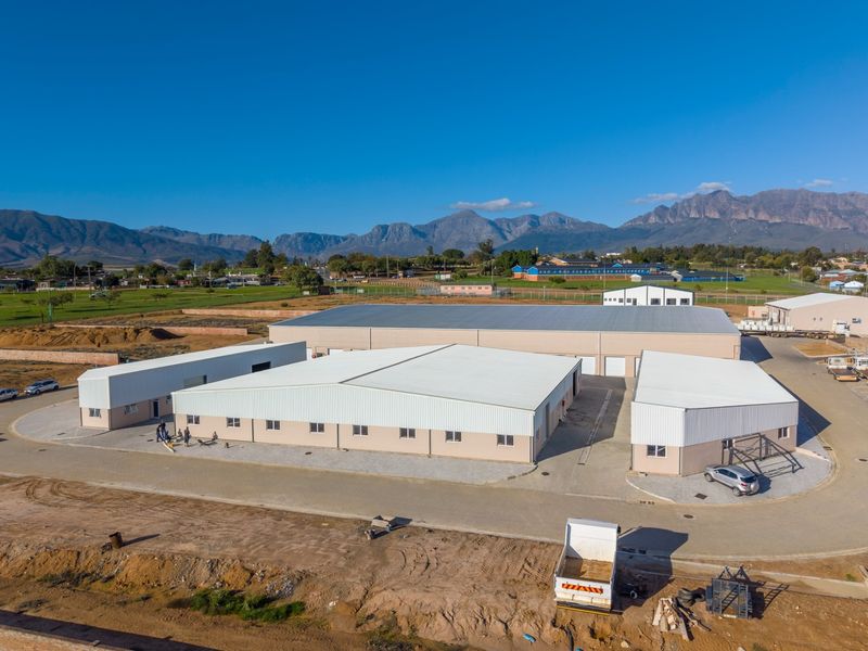Aerial view of a modern industrial warehouse complex featuring multiple metal-roofed buildings, paved access roads, and surrounding open land with mountain views.