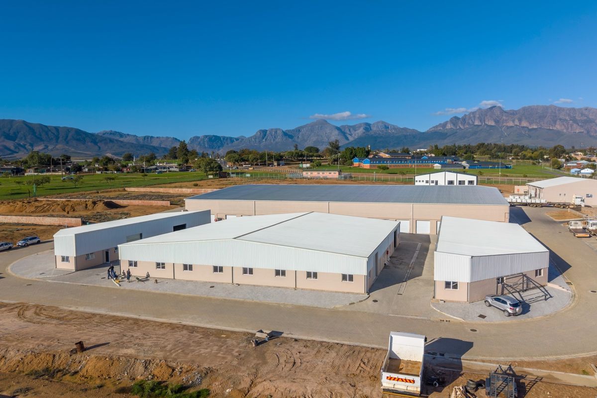 Aerial view of a modern industrial warehouse complex featuring multiple metal-roofed buildings, paved access roads, and surrounding open land with mountain views.
