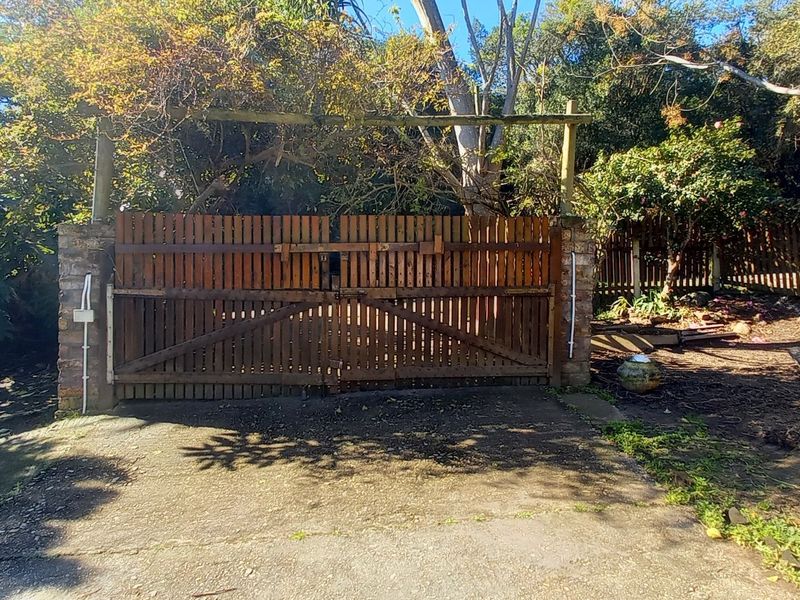 Front entrance featuring a timber driveway gate set between stone pillars, leading to a concrete driveway surrounded by mature trees and fencing.