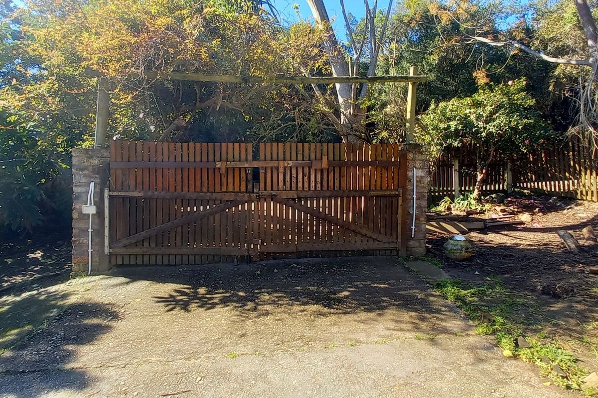 Front entrance featuring a timber driveway gate set between stone pillars, leading to a concrete driveway surrounded by mature trees and fencing.