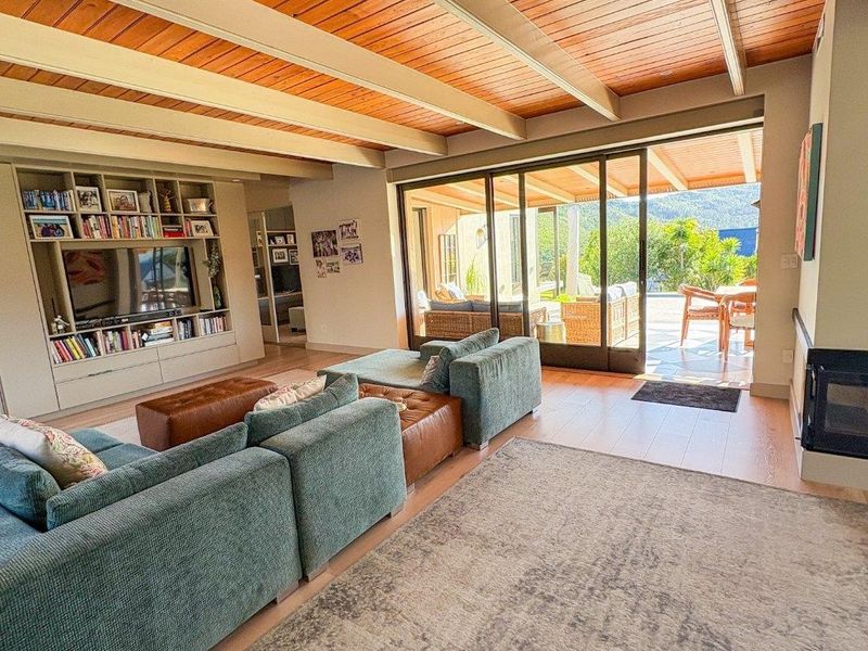 Spacious living room featuring a warm timber-clad vaulted ceiling with exposed beams, built-in shelving, a fireplace, and seamless access to an outdoor patio.