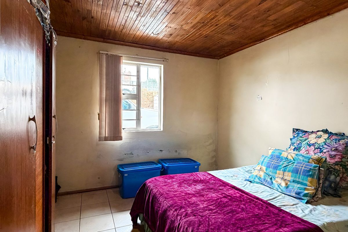 A bedroom featuring a timber ceiling, tiled flooring, and an aluminium window frame, showing signs of wear on the wall surfaces.