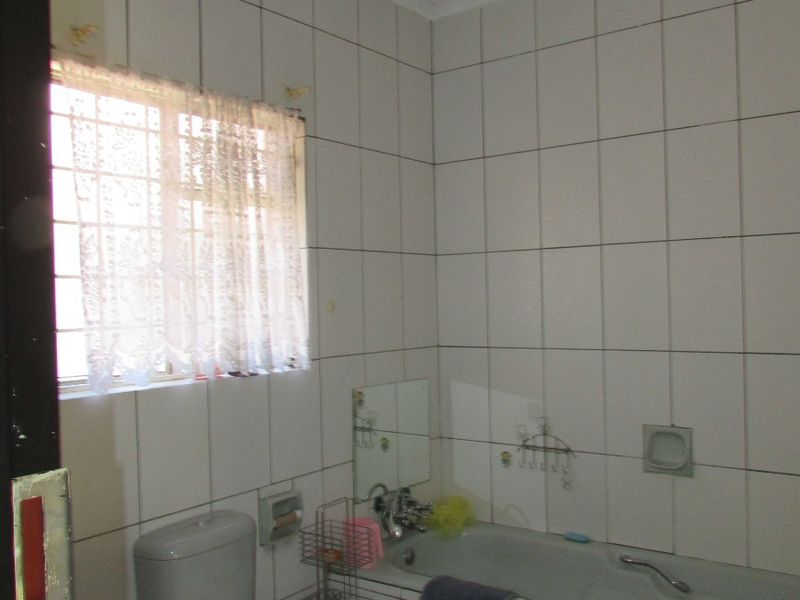 A functional bathroom featuring a built-in bathtub, pedestal basin, and toilet with full-height wall tiling and security bars on the window.