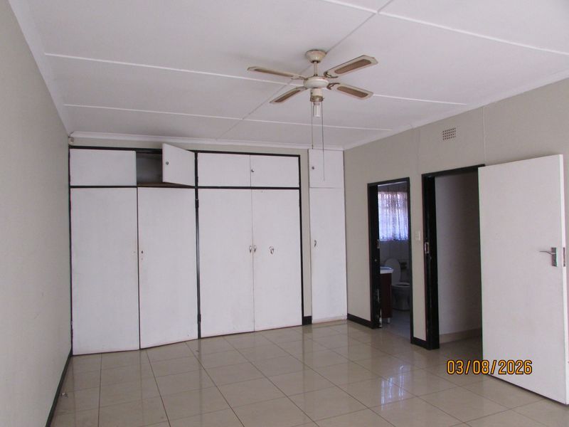 A spacious bedroom featuring tiled flooring, a ceiling fan, and extensive built-in cupboard storage with direct access to an adjoining bathroom.