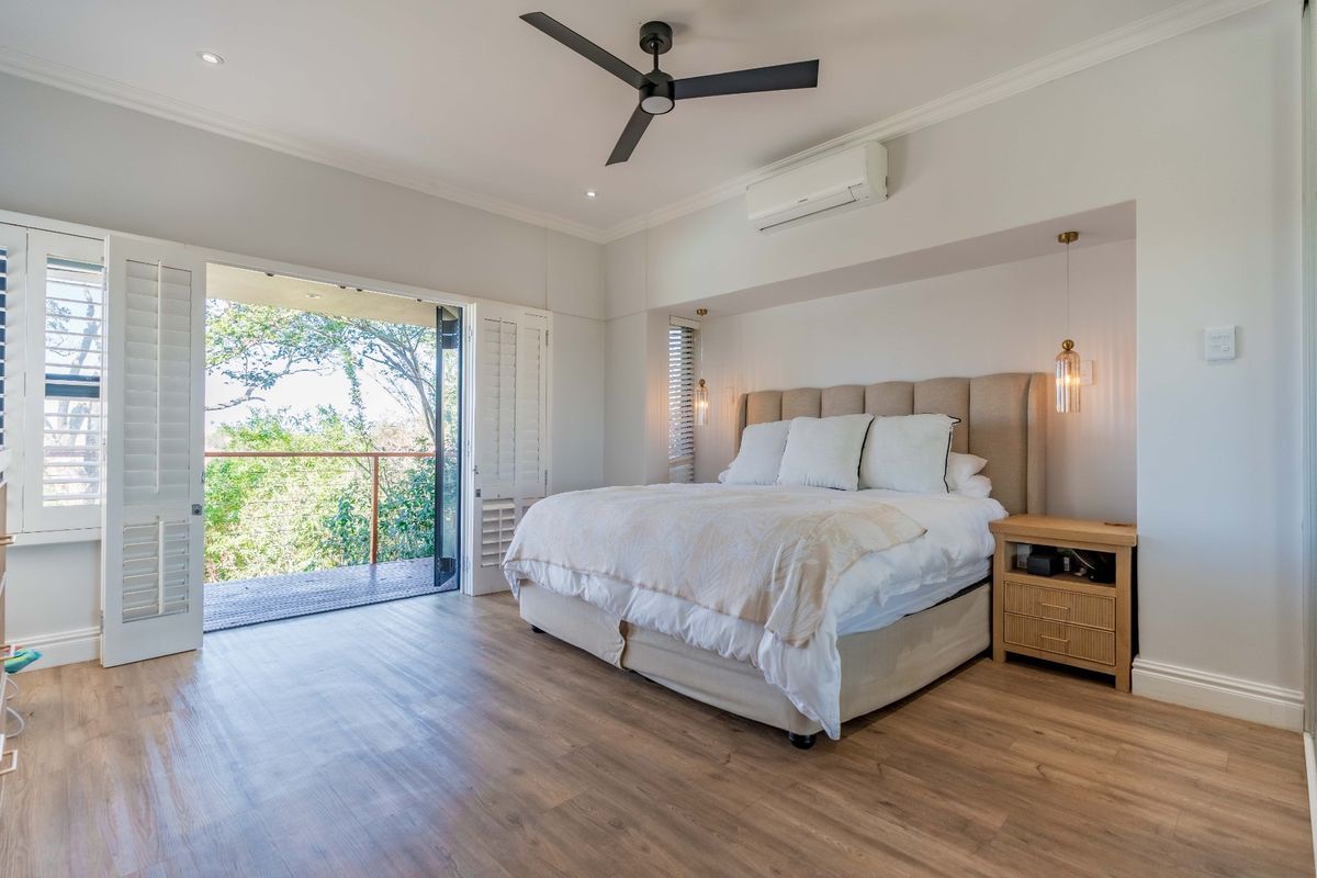Spacious bedroom featuring modern laminate flooring, a ceiling fan, split-system air conditioning, and direct access to a private balcony through plantation shutters.