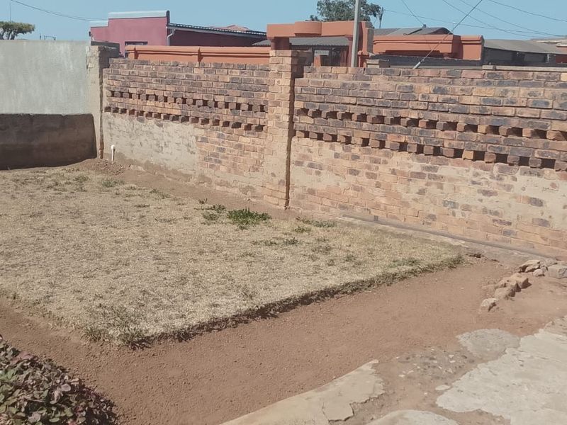 A modest residential garden area featuring a brick boundary wall, metal gate, and a small lawn section requiring landscaping and maintenance.