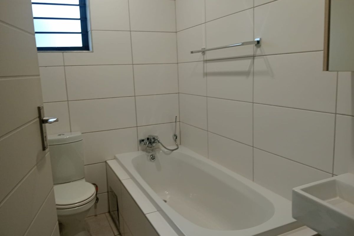 A clean, modern bathroom featuring white wall tiles, a built-in bathtub, a toilet, and a louvered window for ventilation.