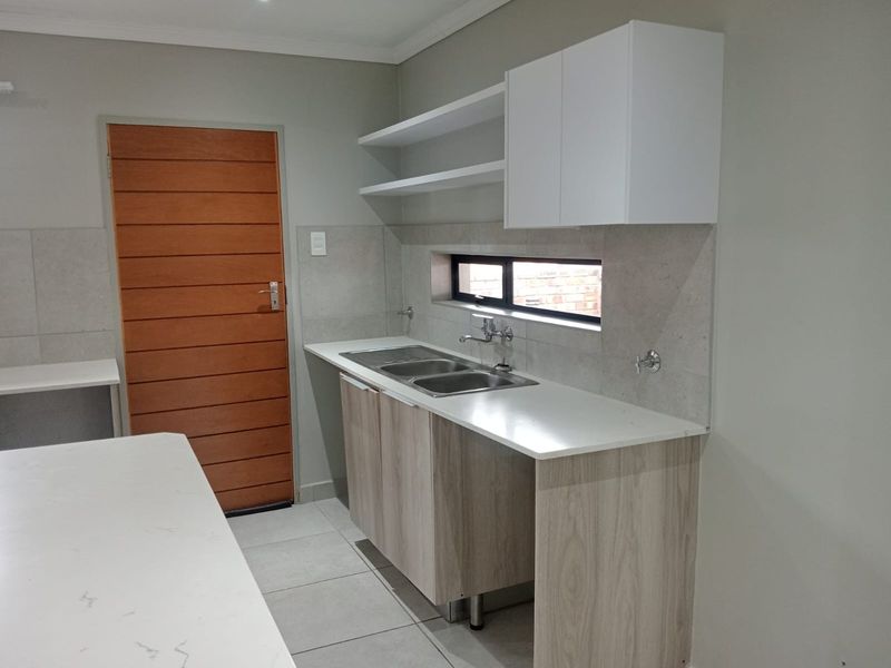 A modern, clean kitchen area featuring light wood-finish cabinetry, stone countertops, a double sink, and neutral tiled flooring.