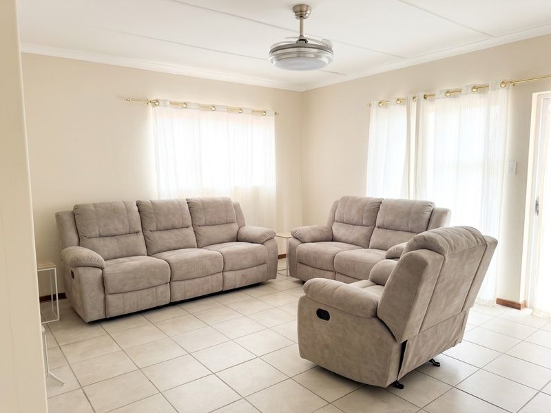 A bright and spacious living room featuring neutral tiled flooring, a modern ceiling fan, and large windows with curtain rods.