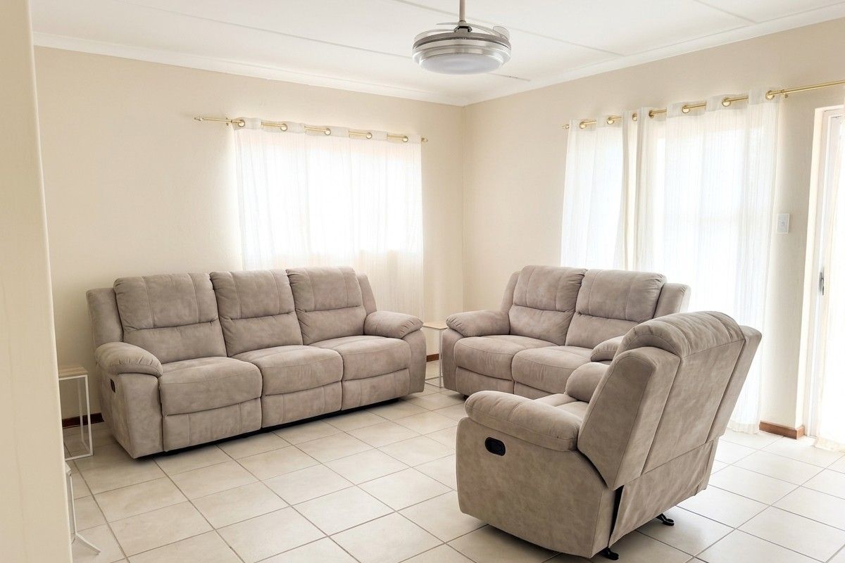 A bright and spacious living room featuring neutral tiled flooring, a modern ceiling fan, and large windows with curtain rods.