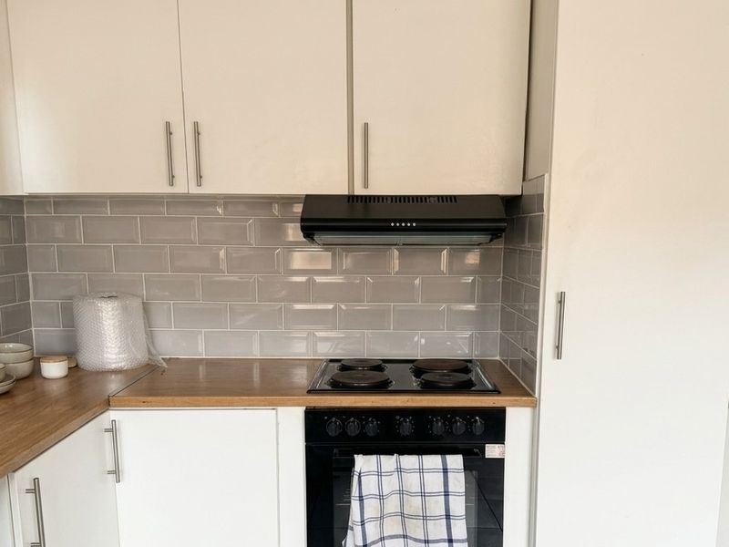 A clean, modern kitchen area featuring white cabinetry, a grey subway tile backsplash, wood-look countertops, and integrated electric cooking appliances.