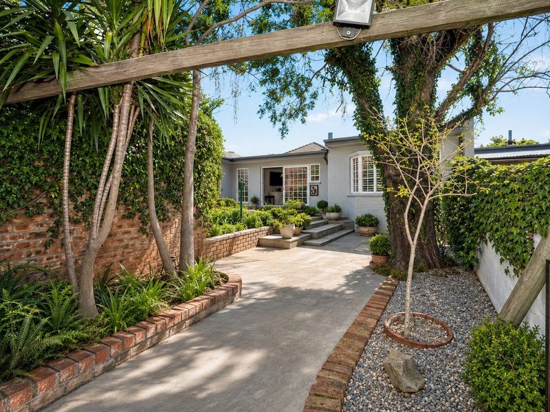 A well-maintained rear courtyard featuring concrete paving, brick retaining walls, mature trees, and direct access to the home through sliding glass doors.