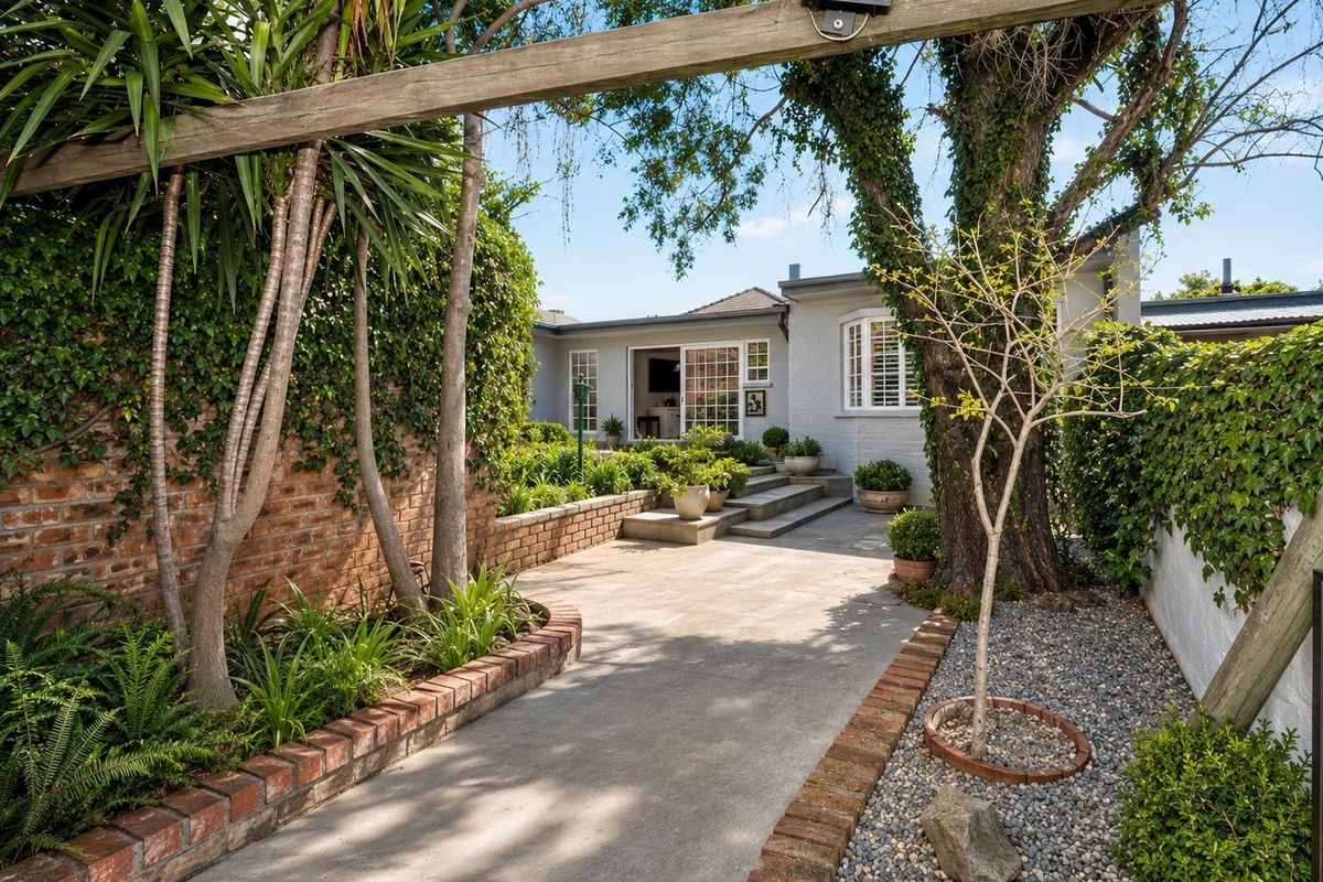 A well-maintained rear courtyard featuring concrete paving, brick retaining walls, mature trees, and direct access to the home through sliding glass doors.