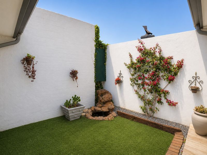 A private, low-maintenance courtyard featuring artificial turf, a stone water feature, and decorative climbing bougainvillea against clean white rendered boundary walls.
