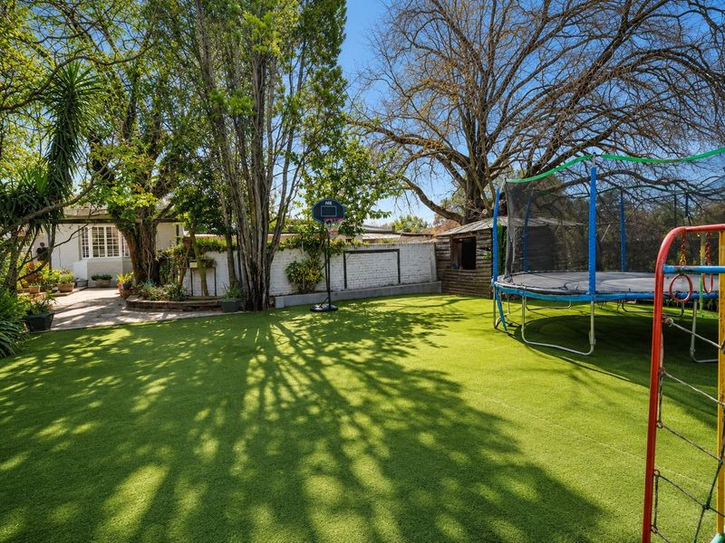 A spacious, well-maintained backyard featuring artificial turf, mature trees, a paved patio area, and a secure boundary wall.