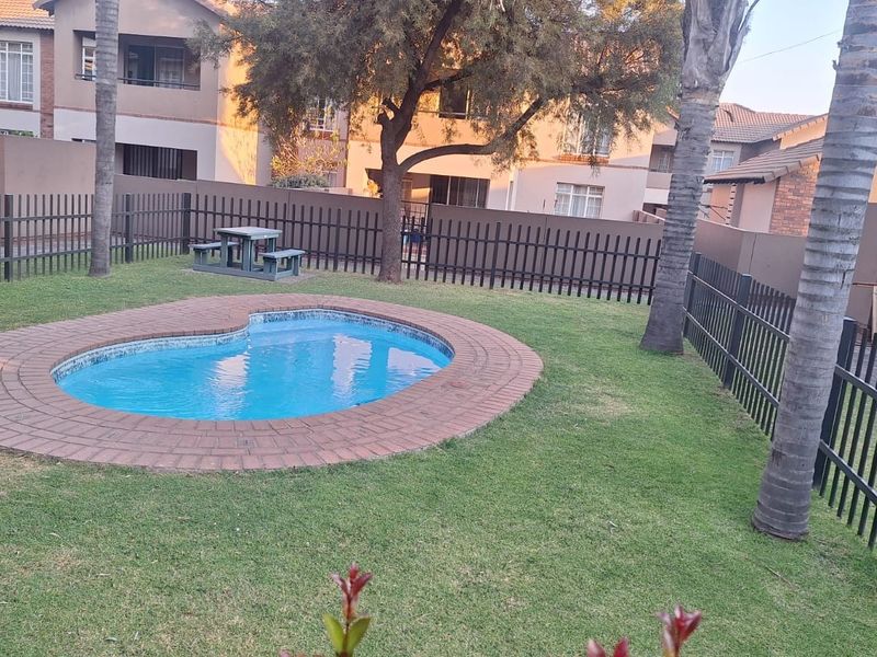 A communal outdoor pool area featuring a kidney-shaped swimming pool with brick coping, surrounded by a well-maintained lawn and secure metal perimeter fencing.
