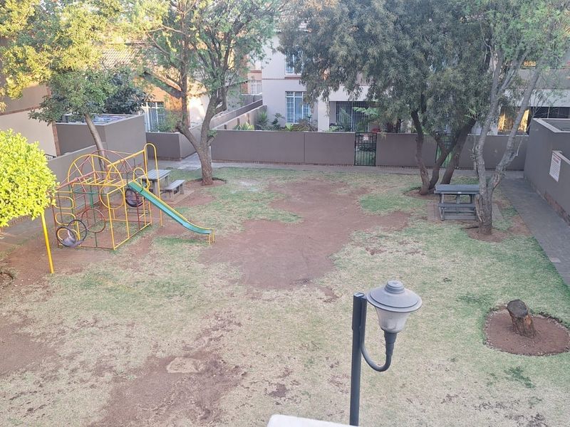 Communal garden area featuring a playground, paved walkways, perimeter walling, and mature trees within the apartment complex.