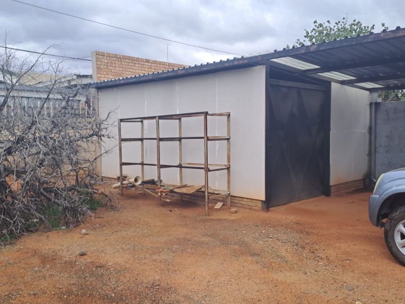 A detached garage structure featuring a metal door and an attached carport, situated on an unpaved lot with perimeter boundary walls.