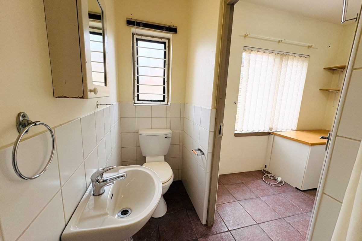 A functional bathroom featuring a wall-mounted basin, toilet, and tiled splashback, adjacent to a small utility or office space with tiled flooring.