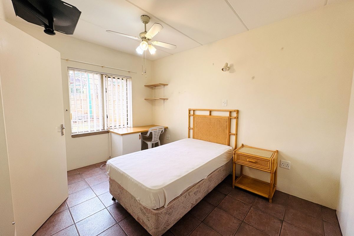 A functional bedroom featuring tiled flooring, a ceiling fan, a built-in desk, and security bars on the window.