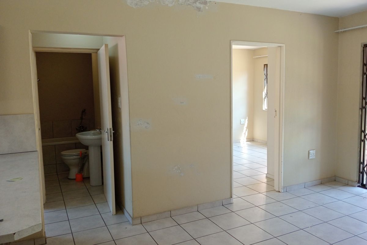An empty living area with tiled flooring, showing access to a bathroom and adjacent room, requiring cosmetic repairs to walls and ceiling.
