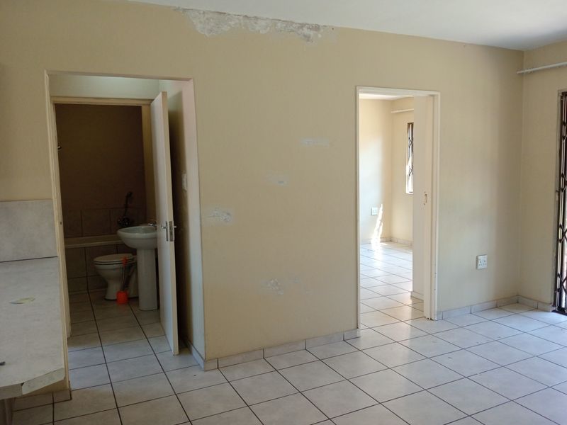 An empty living area with tiled flooring, showing access to a bathroom and adjacent room, requiring cosmetic repairs to walls and ceiling.