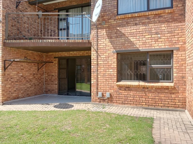 Rear view of a face brick apartment building featuring a ground floor paved patio, sliding door access, and an upper-level balcony with metal railings.
