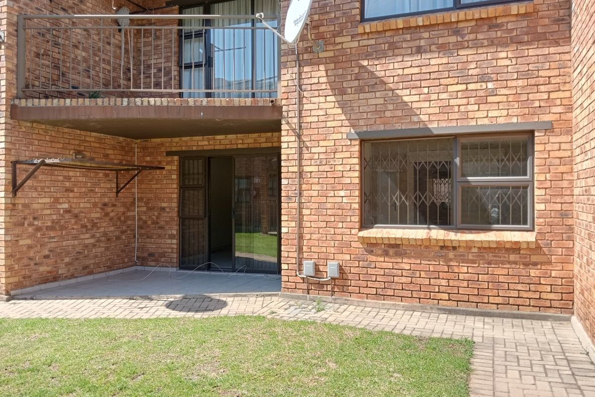 Rear view of a face brick apartment building featuring a ground floor paved patio, sliding door access, and an upper-level balcony with metal railings.
