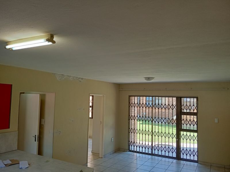 An open-plan living area featuring tiled flooring, a security gate leading to the exterior, and visible wall surface damage requiring maintenance.