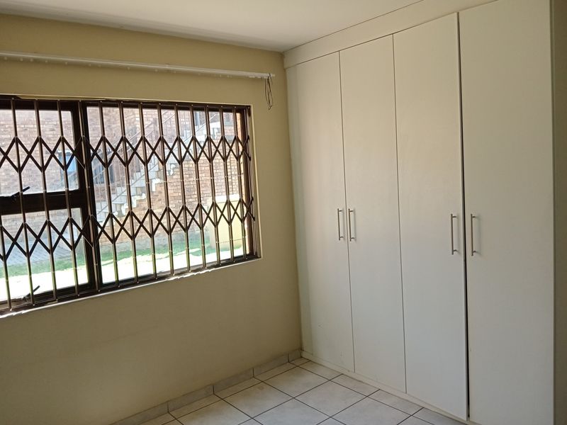 A clean, empty bedroom featuring neutral tiled flooring, ample built-in cupboard storage, and a window fitted with a security gate.