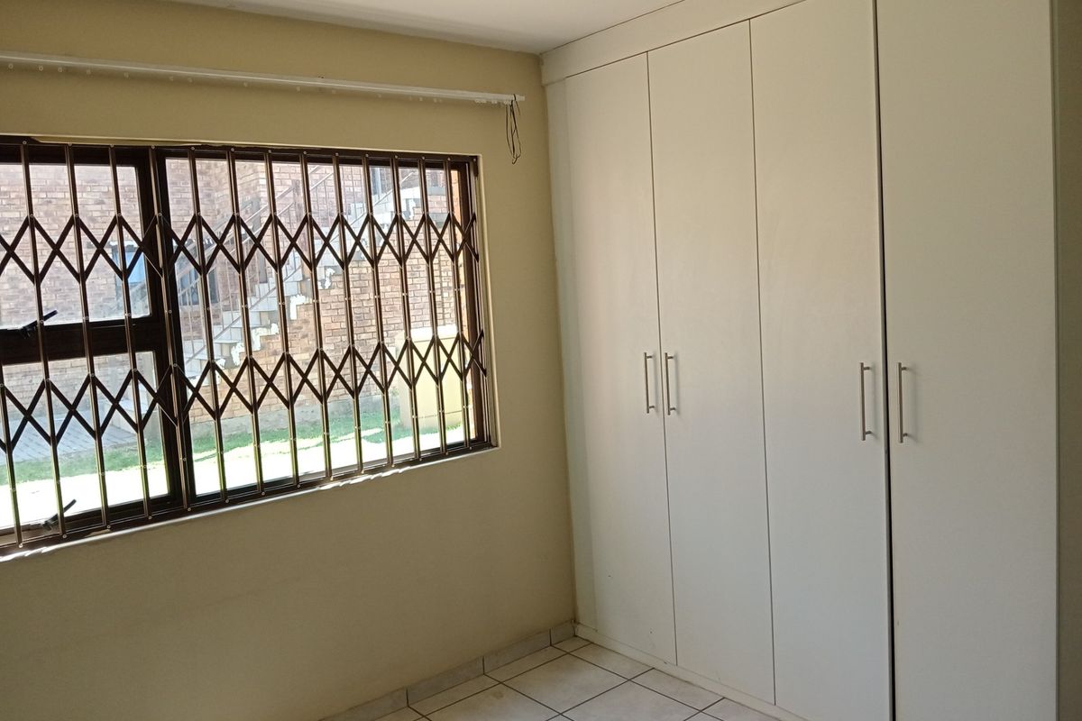A clean, empty bedroom featuring neutral tiled flooring, ample built-in cupboard storage, and a window fitted with a security gate.