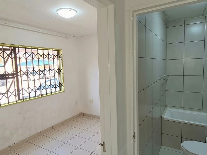 A view from the hallway looking into an empty bedroom with security bars and a tiled bathroom featuring a bathtub, toilet, and basin.