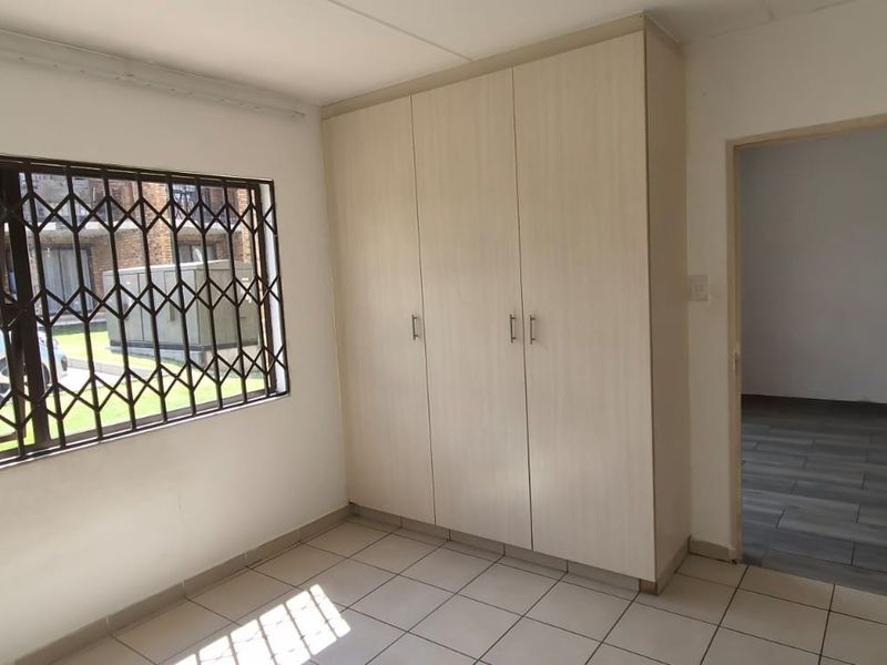 A bright, empty bedroom featuring built-in cupboards, tiled flooring, and a window fitted with a security gate.