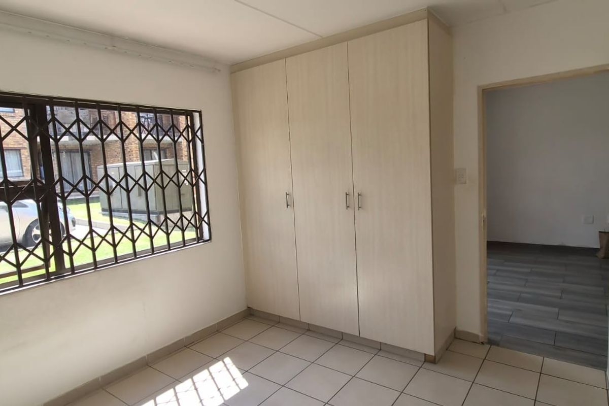 A bright, empty bedroom featuring built-in cupboards, tiled flooring, and a window fitted with a security gate.