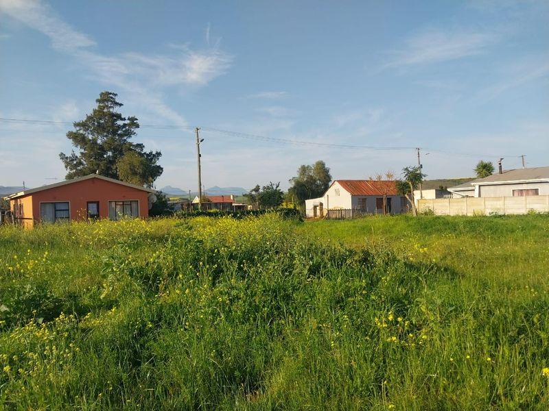 A view of a vacant residential plot featuring natural grassland, with neighboring houses visible in the background.