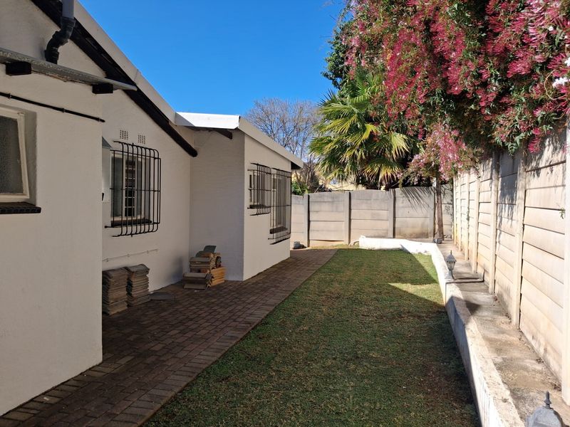 A rear exterior view of the property featuring a paved walkway, a small lawn, and a secure boundary wall with established garden greenery.