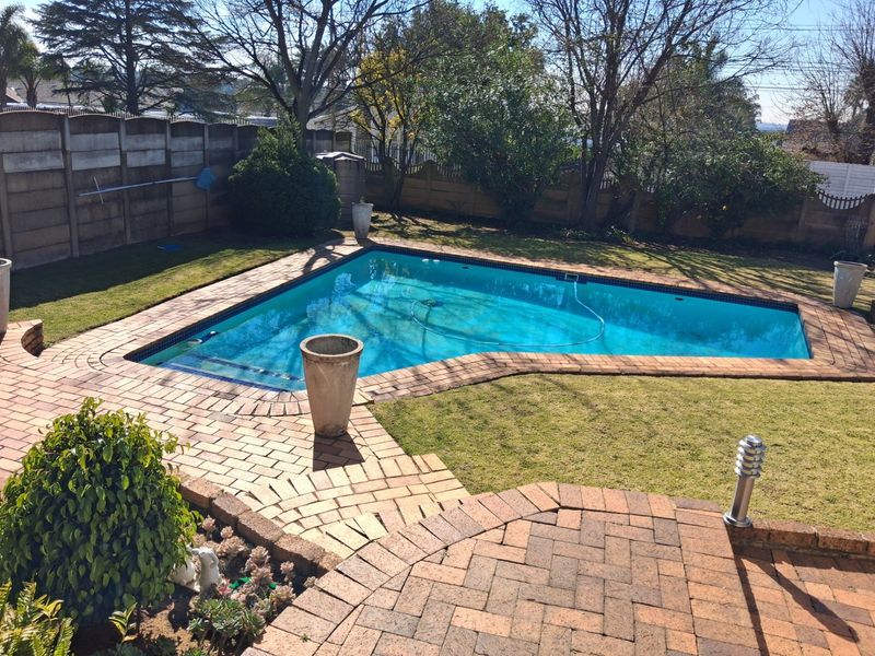 A well-maintained outdoor pool area featuring a swimming pool, brick paved surround, and established garden landscaping with perimeter walling.