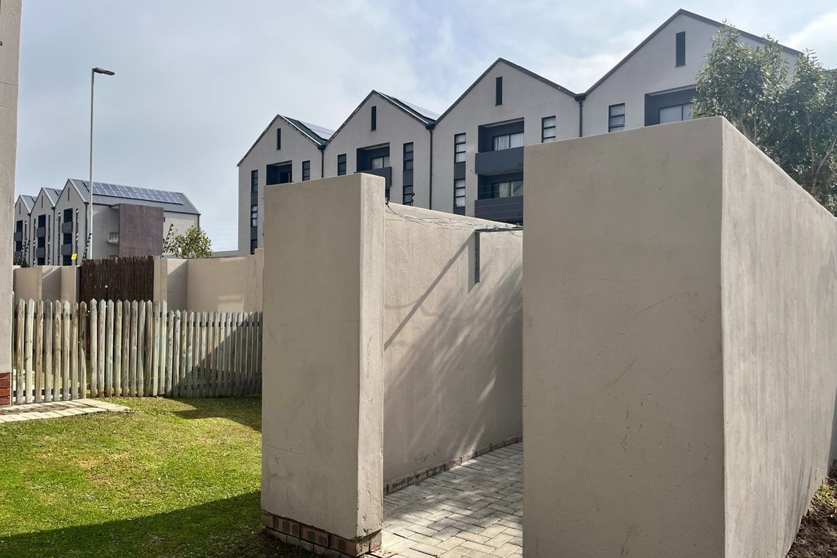 A rear exterior view of an apartment complex featuring a private paved walkway, boundary walls, and a small lawn area.