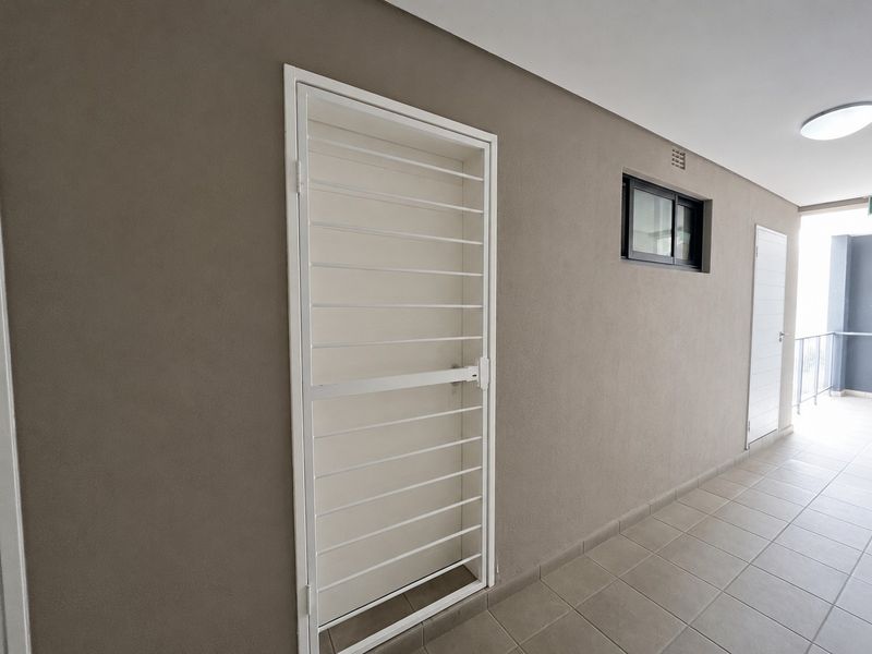 A clean, well-maintained apartment hallway featuring tiled flooring, a white security gate, and a small sliding window.
