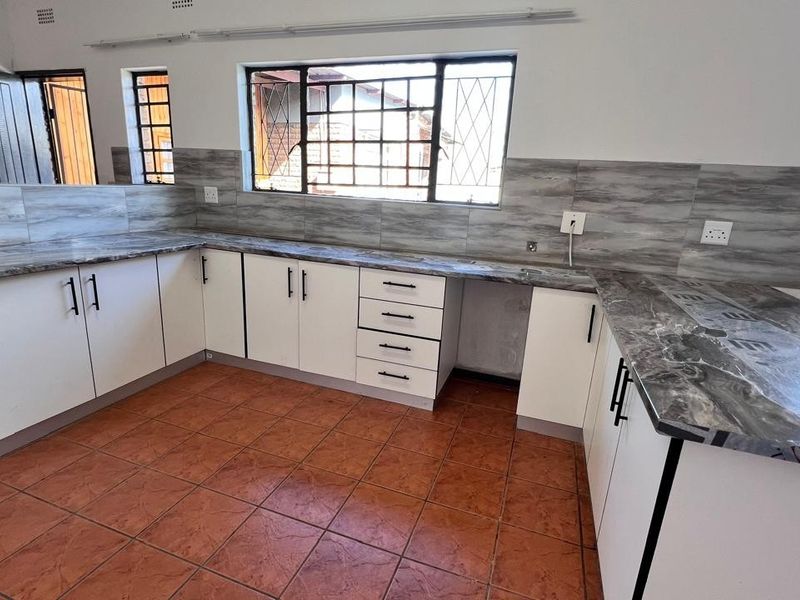 A clean, empty kitchen featuring modern white cabinetry, marble-effect laminate countertops, tiled backsplash, and terracotta-style floor tiles with security bars on the windows.