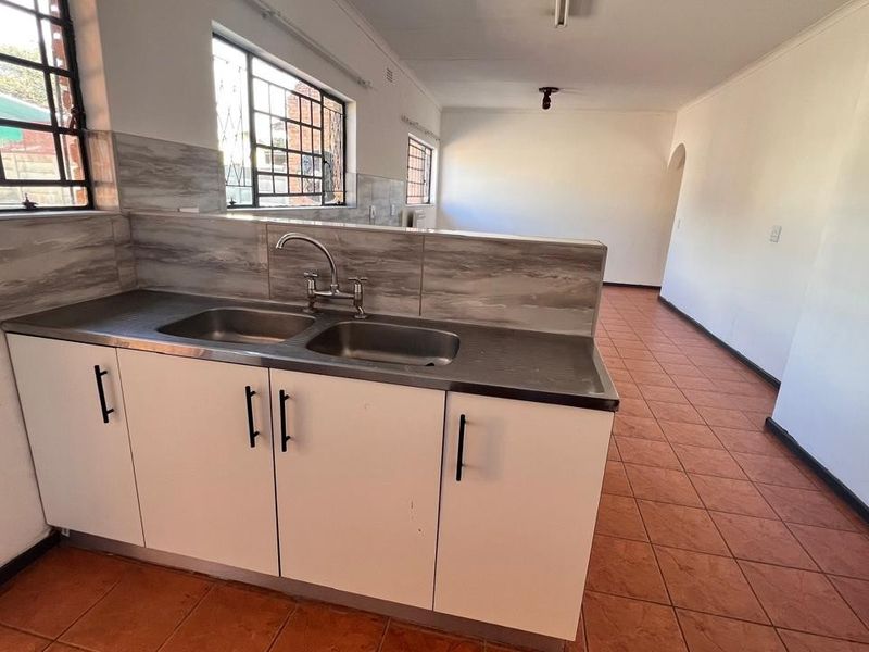 A functional kitchen area featuring base cabinetry, a double sink, tiled backsplash, and terracotta floor tiles, leading into an open-plan living space.