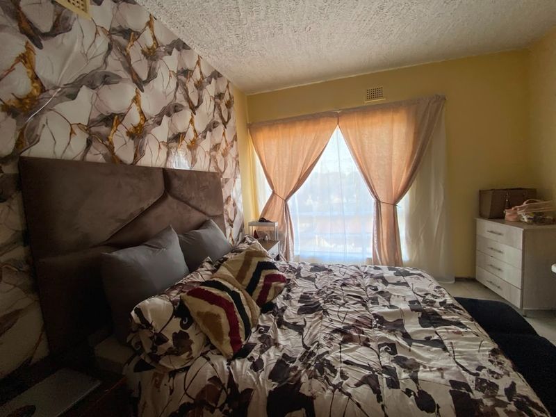 A bedroom featuring a textured ceiling, ventilation grille, and large window with curtains, providing natural light to the space.