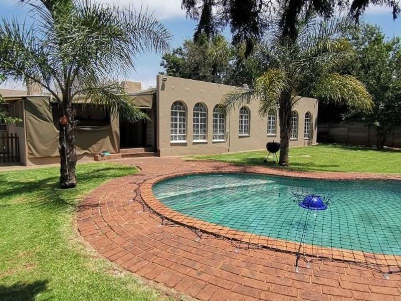 Rear exterior view featuring a swimming pool with safety net, brick paved surround, landscaped lawn, and a covered patio area with arched windows.