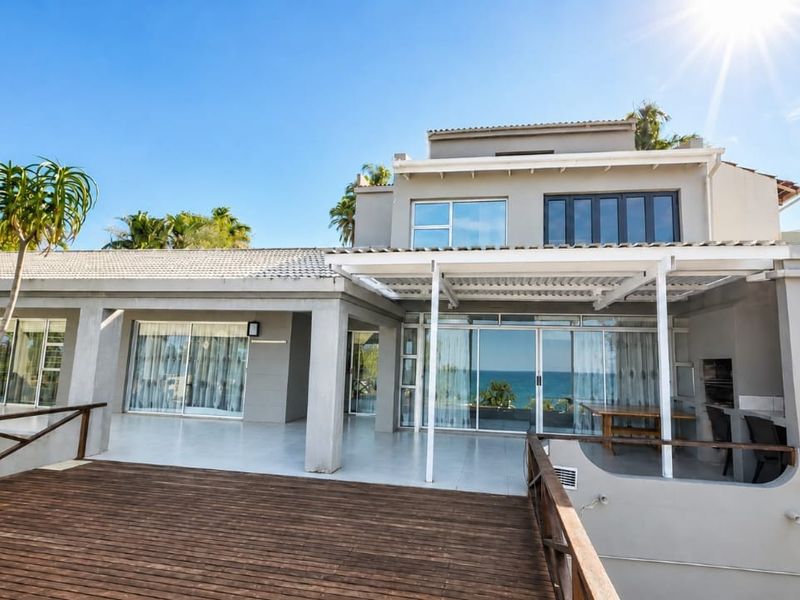 Rear elevation featuring a spacious wooden deck, covered patio area with built-in braai, and large sliding doors overlooking the ocean.