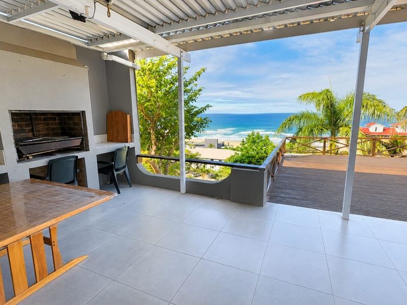 Covered patio featuring a built-in braai, tiled flooring, and an adjoining timber deck with expansive ocean views.
