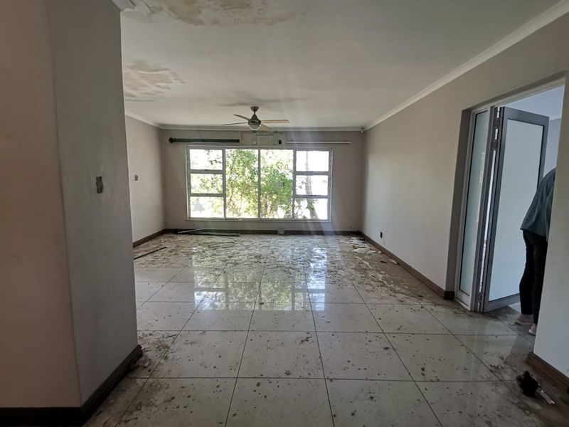 An empty living room featuring large windows, a ceiling fan, and tiled flooring, currently showing signs of ceiling water damage and floor staining.